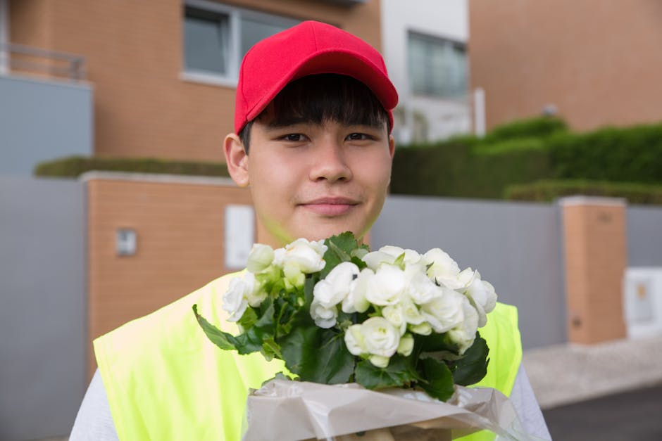 A young male flower delivery courier wearing a red cap and high-visibility yellow vest, holding a bouquet of white roses wrapped in white paper, standing outdoors in front of modern residential buildi
