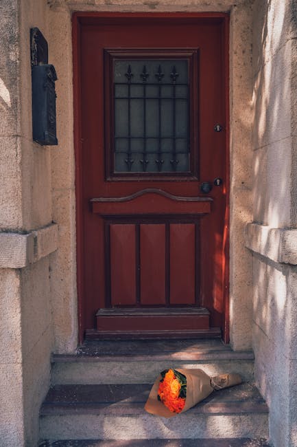 A bouquet of fresh orange and yellow flowers, wrapped in brown paper with a natural twine ribbon, resting on stone steps in front of a red front door with black wrought iron detailing. The flowers inc