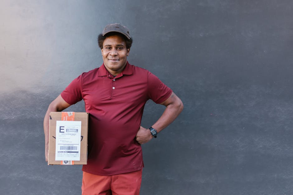 A smiling delivery person with medium-dark skin tone, wearing a maroon polo shirt, matching shorts, and a grey cap, stands against a plain dark grey wall. They are holding a small cardboard parcel wit