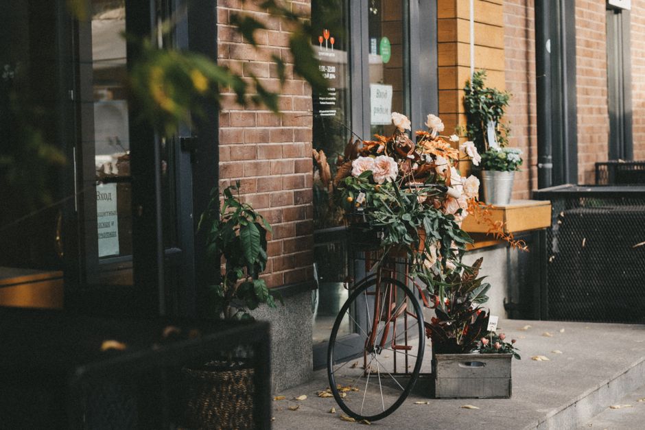 A vintage-style bicycle with a rust-colored frame is parked outside a flower shop on Old Malden High Street. The bicycle's rear basket is filled with fresh, pastel-colored flowers, including pale pink