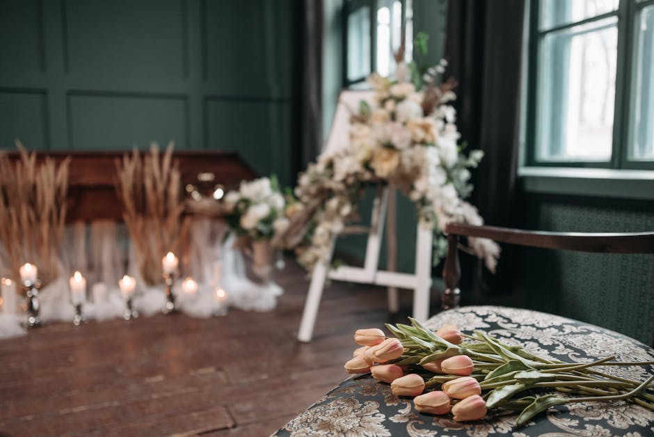 A floral arrangement featuring soft pink tulips with green stems and leaves resting on an ornate upholstered chair in a softly lit room. In the background, there are additional white and cream flowers