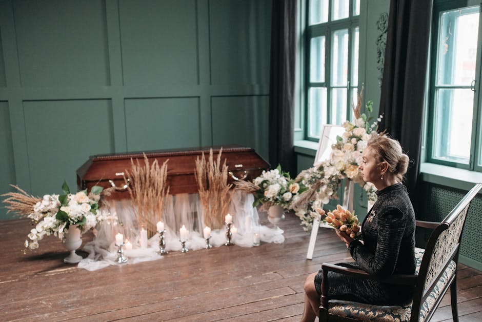 A woman seated on an ornate wooden chair, holding a bouquet of soft peach and cream flowers, faces a floral arrangement composed of white roses, peonies, and greenery, set in a white vase. The arrange