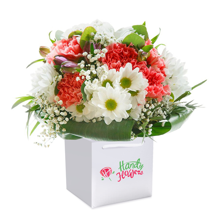 Exquisitely arranged in a sleek, square white gift bag and isolated on a bright white background, this floral bouquet showcases a vivid harmony of colours and textures. The lush focal point is formed by layers of soft coral, peach, and radiant red carnations, their abundant, ruffled petals bringing a sense of festivity and depth. Among them, crisp white daisies with lively yellow-green centres add bursts of freshness, reminiscent of wildflowers glimpsed in Old Malden's leafy parks. Breezy sprays of baby's breath weave between the larger flowers, their petite white blossoms hinting at delicacy and lightness. Toward the left, discreet purple-tinged buds-perhaps alstroemeria, yet to fully open-add nuance and a promise of continued bloom. All are cradled within a generous halo of robust, glossy dark green leaves, providing structure and highlighting each flower's form, not unlike the natural verdure bordering Malden Road. The well-constructed gift bag, complete with white ribbon handles, suggests this arrangement is ready to be gifted at a moment's notice, ideal for special events or everyday tokens of care. The entire composition exudes a sense of professional expertise and local pride, reflecting the quality and attention to detail renowned among florists in Old Malden. Whether sent for a birthday celebration, an anniversary, or simply to bring cheer to a home in or around Blagdon Road, this bouquet offers both visual impact and heartfelt intent-each stem hand-selected to capture a spirit of generosity and seasonal renewal in the heart of the community.