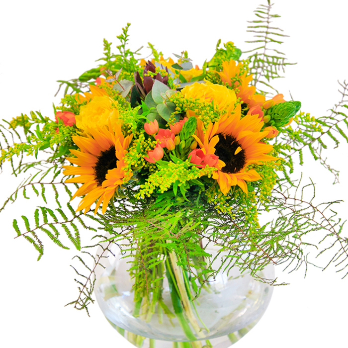 Radiating all the exuberance of a sun-streaked afternoon in Old Malden, this lush arrangement is artfully displayed in a perfectly round, clear glass vase. At its center stand bold, beaming sunflowers-their golden yellow-orange petals flaring out from deep, dark centres, reminiscent of warm fields just beyond Motspur Park. Encircling these sunny giants, tightly whorled yellow garden roses unfurl elegantly, their layered petals glimmering softly in the ambient daylight. Intermixed are sprigs of bright red Hypericum berries that sparkle like scattered jewels amidst a mass of greenery, catching the eye with each glossy curve. The bouquet is exuberantly filled with diverse living textures: arching fronds of soft, feathery fern, airy light green filler lightly dusted with miniature yellow flower heads, and slender, spike-like greens adding subtle dynamism. Bluish eucalyptus leaves peak modestly from behind, injecting the arrangement with a fresh, herbaceous note that cools the vivid yellows and reds. The base of the vase reveals the neatly trimmed stems, their reflections dancing in water to further amplify a sense of vitality and freshness. This composition captures the spirit of a jubilant moment-be it the first sunlit days of the season, a joyful anniversary, or a 'just because' surprise for someone around Old Malden. The naturally harmonious mix of flowers and foliage, paired with the craftsmanship honed by local florists, creates a bouquet that doesn't just brighten rooms but also brings the cheerful optimism of summer wherever it goes.